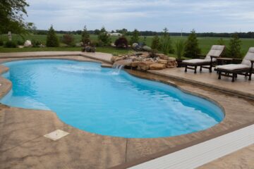 A light-blue freeform inground pool surrounded by light brown stamped concrete with a rock formation waterfall on the right-side length of the pool. Two lawn chairs sit next to the waterfall. The pool patio is surrounded by small trees that wrap around it.