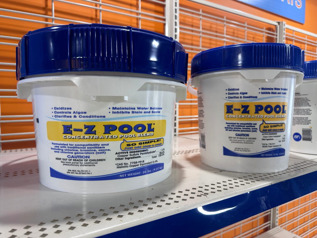Landscape photo of two buckets of E-Z POOL. The bucket on the left is a twenty-pound bucket, and the one on the right is a ten-pound bucket. The buckets are predominantly white with blue lettering and blue lids. The E-Z POOL logos are imposed over a yellow rectangle. The buckets sit atop a white shelf with a blue front-edge in front of an orange wall behind white cage-links.