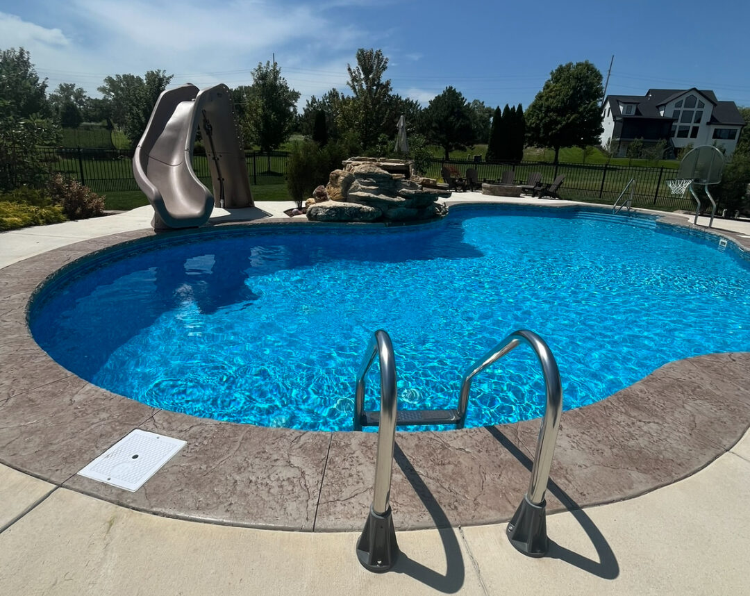 Medium-closeup of a light-blue freeform inground pool with a rock formation waterfall off the middle of the length side in the background of the image. A waterslide is next to the waterfall. Across from the water fall is a basketball hoop. Across from the waterslide is a pool ladder. The pool is surrounded by a stamped concrete border with a larger concrete patio surrounding it. Behind the pool in the background are trees, grass, and a black fence.