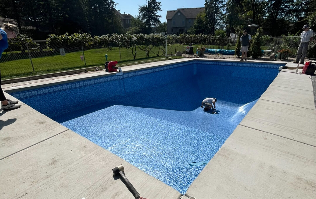 Photo of an empty pool with a new vinyl liner. A man is crouched down in the middle of the deep end. There is a vacuum on the left of the pool sucking the vacuum to the wall. A woman is looking into the pool from the shallow end and two men are looking in from the deep end. The pool is surrounded by brushed concrete and enclosed in a metal fence. 
