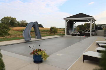 Inground pool covered by a gray automatic pool cover surrounded by a concrete pool deck. The pool features a slide and basketball hoop. A gazebo can be seen in the background close to the edge of the pool. Beyond the yard is grass and a cornfield.
