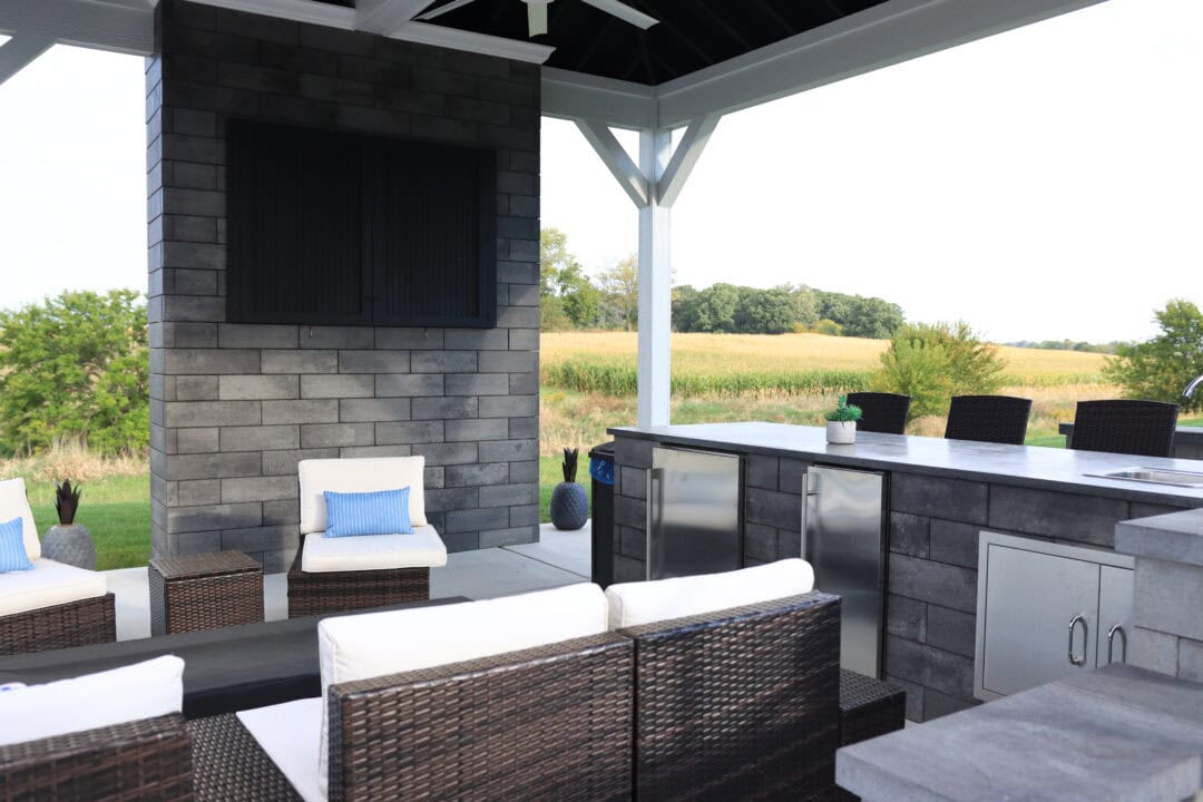 Landscape photo taken from under a residential gazebo. The gazebo features a large brick wall left of center of the image with a large, black, rectangle at the top in the middle. In front of the wall sits five brown and white chairs surrounding a black table. Surrounding the chairs in the bottom right corner of the image is a wrap around counter with a sink, cabinet, and a refrigerator. Three black chairs sit outside the gazebo on the other side of the counter. Grass, trees, and cornfields are visible in the background beyond the gazebo.