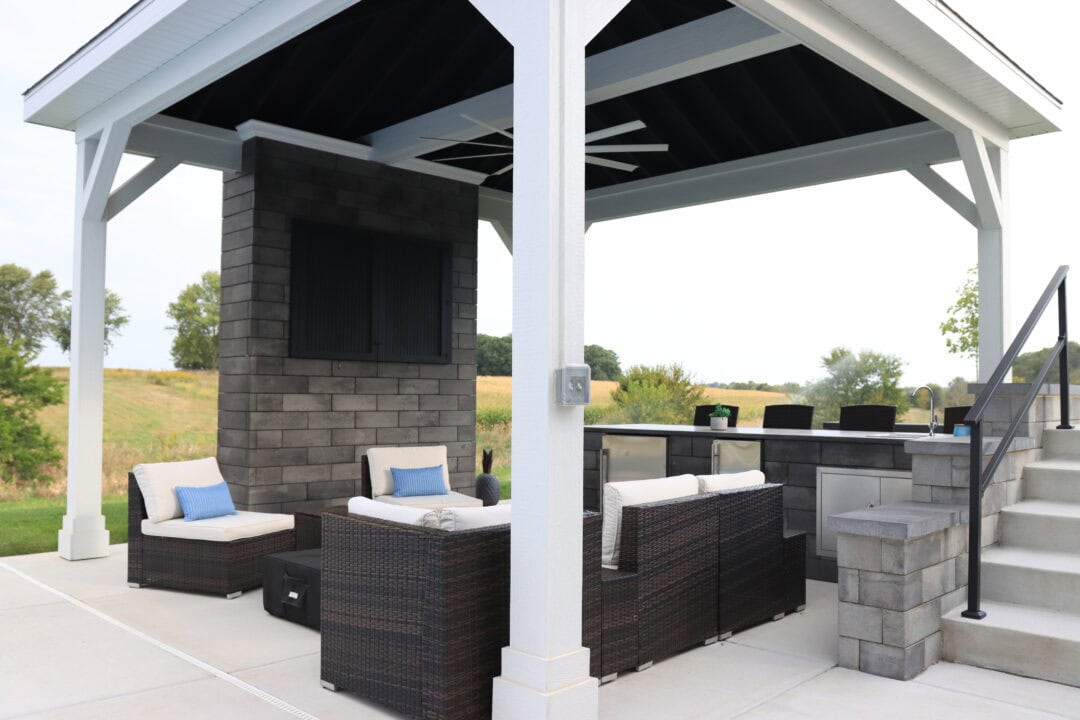 Landscape photo of a gazebo, under which contains a wall, multiple lounge chairs, and an outdoor kitchen. A set of concrete stairs sit next to the gazebo on the far right-side of the image. 