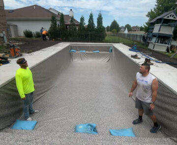 Portrait photo taken from a head-angle pointed down looking at the shallow-end of an empty vinyl liner pool. Two men stand in the shallow end in front of three light-blue bags pressed against the base of the stairs. The liner is a light-gray color. The pool is surrounded by a light-gray concrete patio. The patio is surrounded by dirt in an open yard. In the background are more houses and trees line the width of the deep end in the background. The top third of the image is the sky which is mostly clear with some clouds.