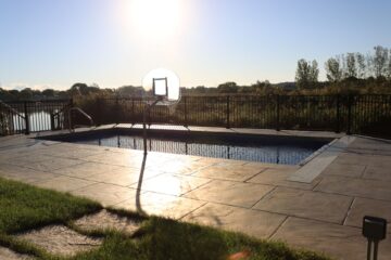 Photo of an inground pool surrounded by a large, tan, concrete patio. The sun can be seen behind the pool in the background. It is reflecting brightly off the concrete in front of the pool closest to camera.