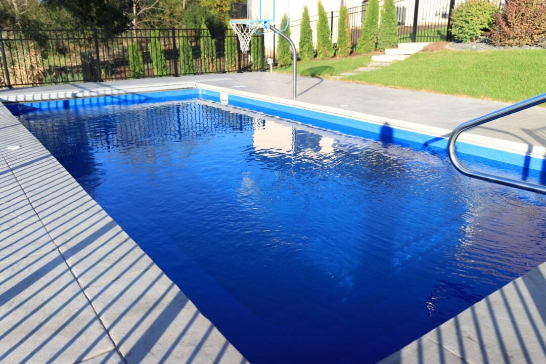 Photo of a blue inground pool taken from the shallow end. The pool features a walk in handrail, a basketball hoop, a retaining wall and textured concrete coping.