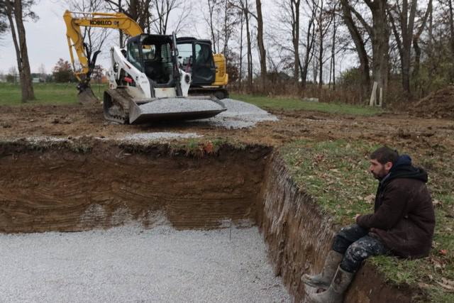 Pool hole being filled with gravel by an excavator. man sitting on pool hole edge.