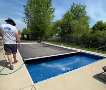 Square photo of a man standing next to an inground pool that is 1/4 open from a gray automatic pool cover. The pool is surrounded by tan concrete. The pool extends from the bottom right corner of the image up to the top left. In the background surrounding the pool patio is grass and trees.
