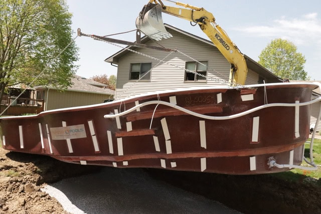 A crane lowering a fiberglass pool into an excavated hole.