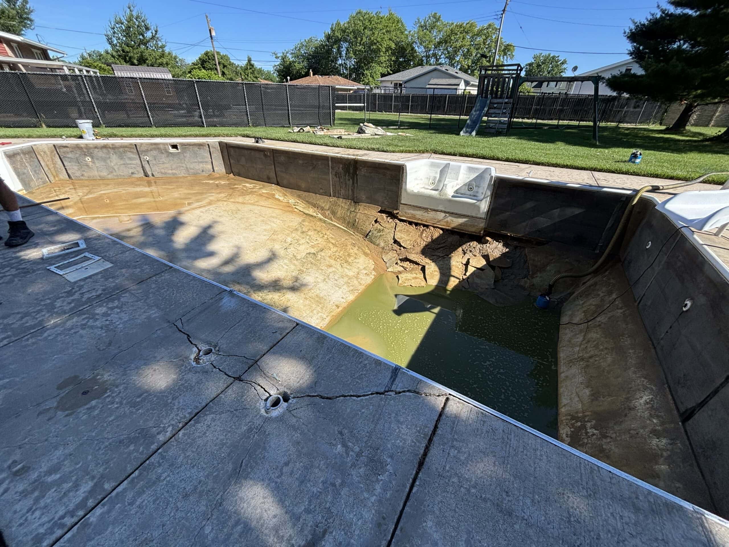 Landscape photo taken of a drained, degraded inground pool hole with steel walls and a vermiculite floor. The concrete surrounding the pool structure can be seen cracked along the bottom of the image. Grass surrounds the patio in the background. A brown fence surrounds the grass. The tops of additional houses can be seen in the background.