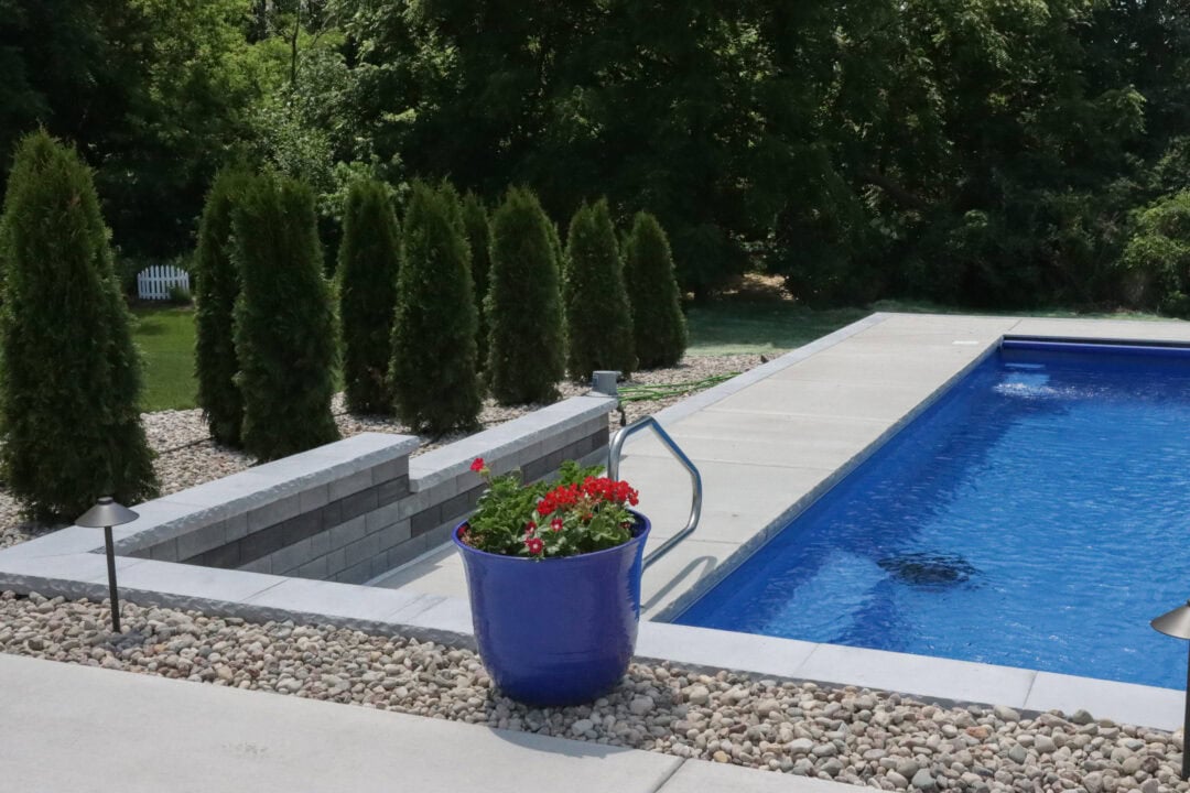 Landscape photo taken of a portion of a concrete pool patio in a backyard. The patio extends at an angle from the bottom left corner of the image to the upper right quarter of the image before extending right to the end of the image. At the bottom of the image is a thin rock bed against the edge of concrete that drops down to the pool patio via retaining wall on the left. A blue, rectangular inground pool can be seen on the left side of the image cut off by the edge. Small evergreen trees line the length side of the patio behind the retaining wall. The background is comprised of grass and tall, thick trees.