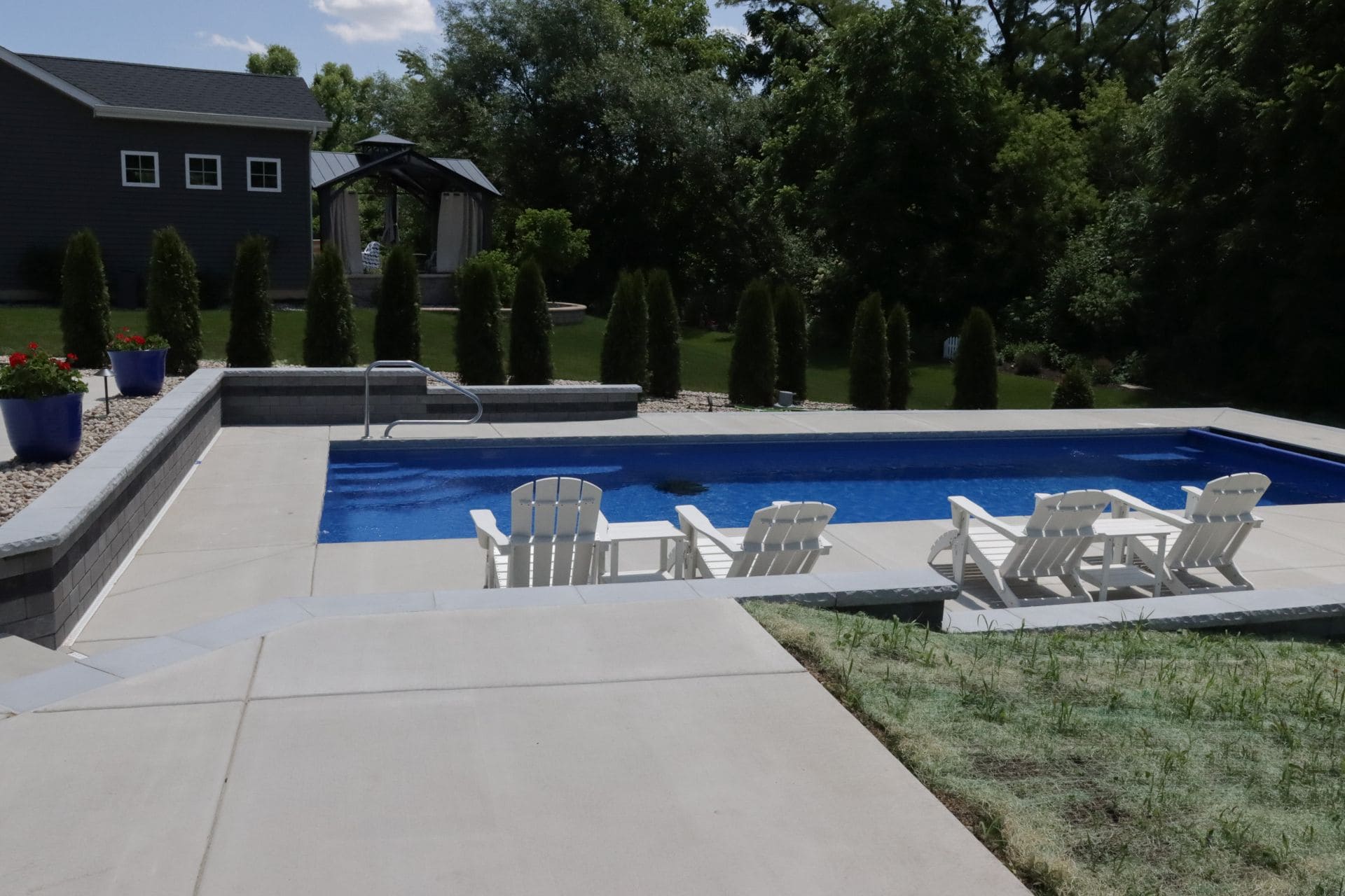 Phot of an inground pool surrounded by a large concrete pool deck. The pool features a retaining wall around the shallow-end width of the pool on the left-side of the image.