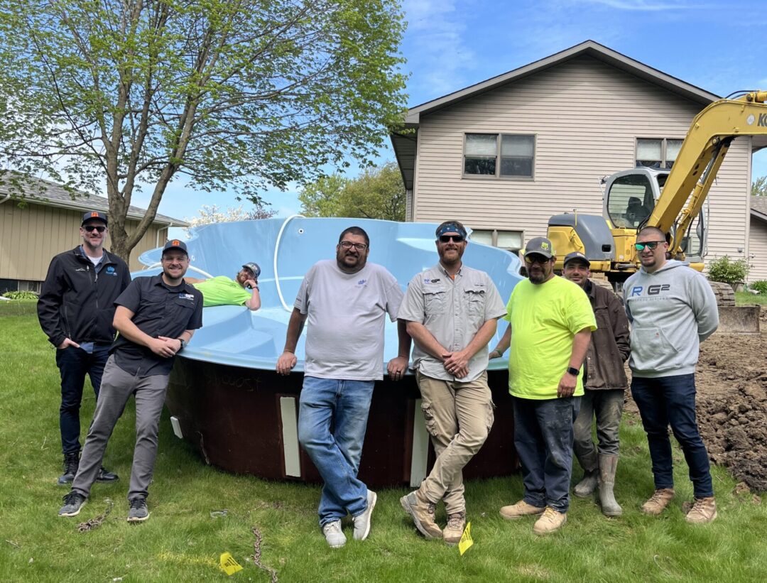 Fiberglass pool sitting on grass in a backyard next to a hole and multiple men standing in front of it.