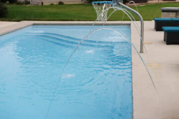 Angled-down photo of the shallow end of a light-blue fiberglass inground pool. The pool features a tanning ledge, and a basketball hoop and three deck jets on the side the photo was taken from.