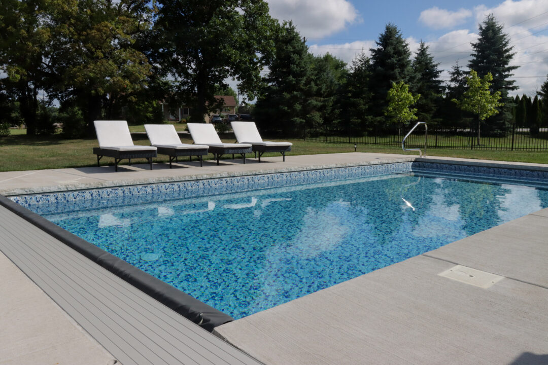 Landscape photo of an inground vinyl liner pool surrounded by a concrete deck. The pool features an automatic cover with a inline box that runs across the bottom left corner of the image. The pool extends from that bottom corner of the image to the edge of the right side. Four black and white lounge chairs sit outside the pool on the left side of the image. Grass and trees surround the patio in the background.