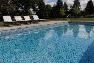 Landscape photo taken at a low angle close to the water level of an inground vinyl liner pool. The pool takes up the bottom three-fourths of the image. Four black and white lounge chairs sit outside the pool on a portion of a concrete pool deck. The pool features an entry handrail, which can be seen near the top right corner of the image. In the background is grass surrounding the patio, and multiple large trees taking up the rest of the background.