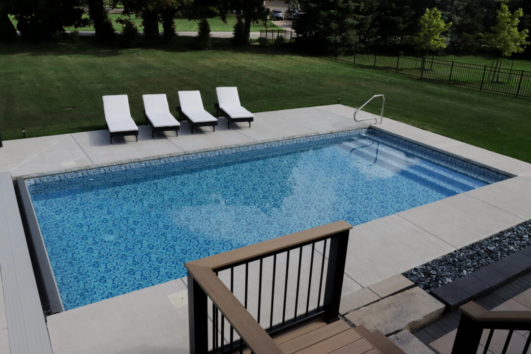 Landscape photo of an inground vinyl liner pool in a backyard. The pool is surrounded by a light-gray concrete patio, upon which sits four black and white beach chairs parallel with the length closest to the grass in the background. Beyond the grass in the background are trees lining the edge of this yard and a neighboring yard on the right. This neighboring yard features a black metal fence stretching along the top right corner of the image. The photo was taken from atop a raised wood deck behind the pool in the foreground. The railing and floor of this deck are visible in the bottom right corner of the image.