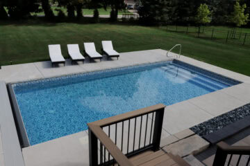 Landscape photo of an inground vinyl liner pool in a backyard. The pool is surrounded by a light-gray concrete patio, upon which sits four black and white beach chairs parallel with the length closest to the grass in the background. Beyond the grass in the background are trees lining the edge of this yard and a neighboring yard on the right. This neighboring yard features a black metal fence stretching along the top right corner of the image. The photo was taken from atop a raised wood deck behind the pool in the foreground. The railing and floor of this deck are visible in the bottom right corner of the image.