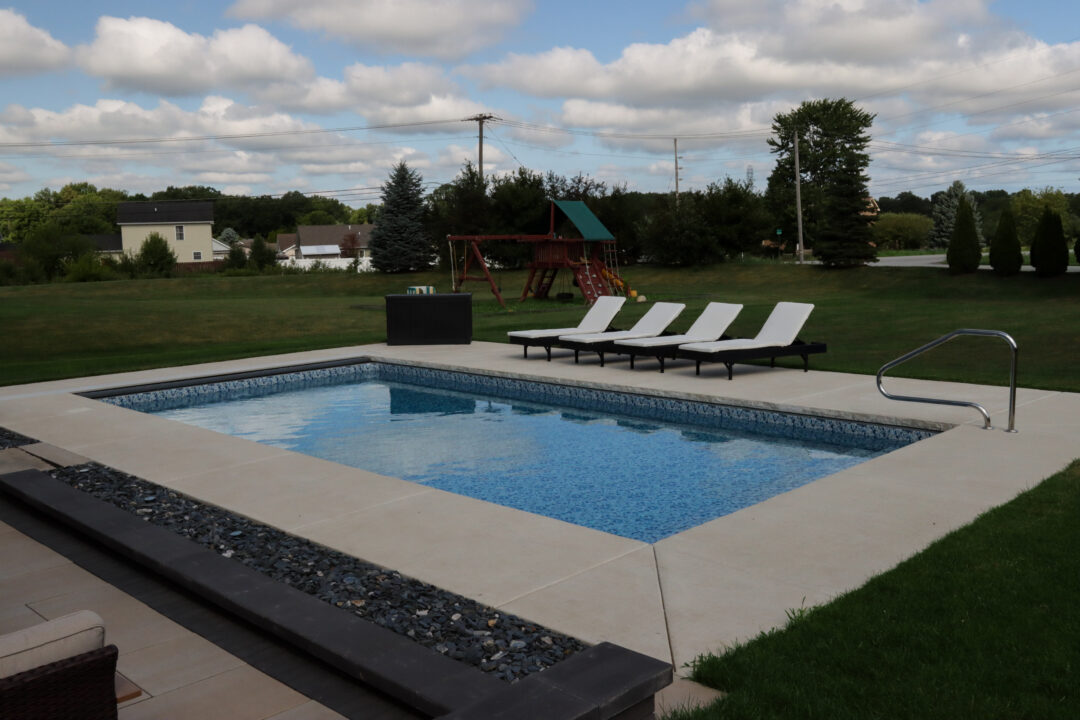 Landscape photo of an inground vinyl liner pool with a handrail in a backyard. The pool is surrounded by a light-gray concrete patio, upon which sits four black and white beach chairs parallel with the length closest to the grass in the background and a rectangular black container next to them in the corner of the patio. On the grass in the background is a brown wood playground set. Additional houses and a road can be seen in the background beyond the yard and trees.