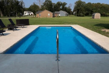 Landscape photo of a blue inground fiberglass pool taken at a low angle from in front of the shallow end directly ahead of a metallic entry handrail. The pool is surrounded by a small concrete patio, atop which on the left sit three black lounge chairs and a large black container at the corner of the patio. Beyond the fence, in the background, is an open grass field. Three houses can be seen in front of trees in the horizon taking up the top fourth of the image.