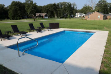 Angled landscape photo of a light blue inground pool surrounded by a light gray concrete patio in an unfenced backyard. The photo is taken at an angle from the right corner of the shallow end, with the pool extending from the bottom left corner to the top right corner. The pool features a metal handrail on the shallow end in the foreground bottom left corner of the image. On the left side of the pool near the top left corner are four black lounge chairs with stands between them, and a container at the end near the edge of the pool and patio. Beyond the pool is a wide open yard, with neighboring houses seen in the background, with trees hanging over them covering the horizon and upper quarter of the image.