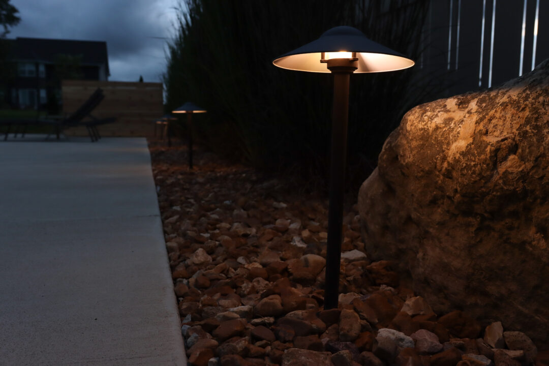 Landscape photo taken at dusk of a stand light and row of stand lights behind it sitting in a rock bed located between a concrete patio on the left and a fence on the right side of the image. Four lights are visible, with one in the foreground focus next to a boulder. Two black lounge chairs are visible left of the lights atop the patio. A house can be seen in shadow in the background.