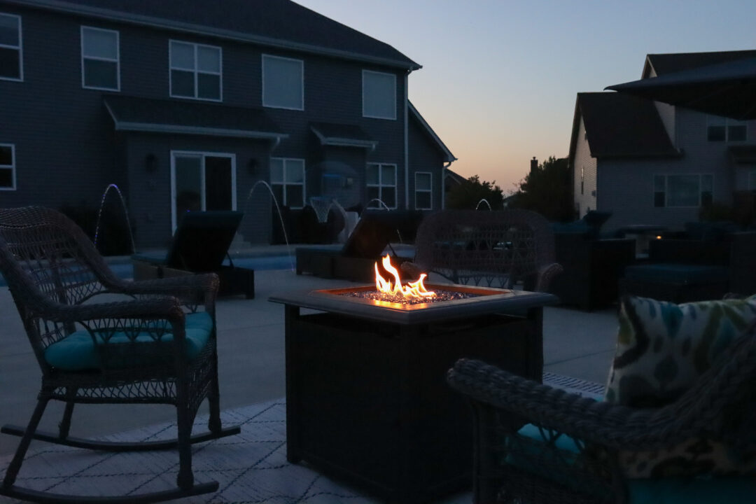 Landscape photo of part of a backyard. A square fire pit sits at the bottom of the image slightly right of center on top of a rug atop a concrete patio. The pit is surrounded by two woven rocking chairs in both bottom corners of the image. Multiple black lounge chairs sitting outside an inground pool can be seen several feet behind the fire pit in front of a dark blue house. A neighboring house can be seen in the upper left corner cut off by the edge of the image. It is dusk.