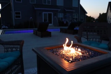 Landscape photo taken from a seated position in front of a lit-up square fire pit sitting atop a concrete patio. The fire pit sits in the bottom right-corner of the image and two cushioned-chairs sit at both corners. In the background is a glowing purple inground pool with four laminars shooting water into the pool and two black lounge chairs sitting outside it several feet behind the fire pit. A house can be seen behind the pool in the top third of the image. A dim sunset sky is visible in the top right corner of the image next to the house.