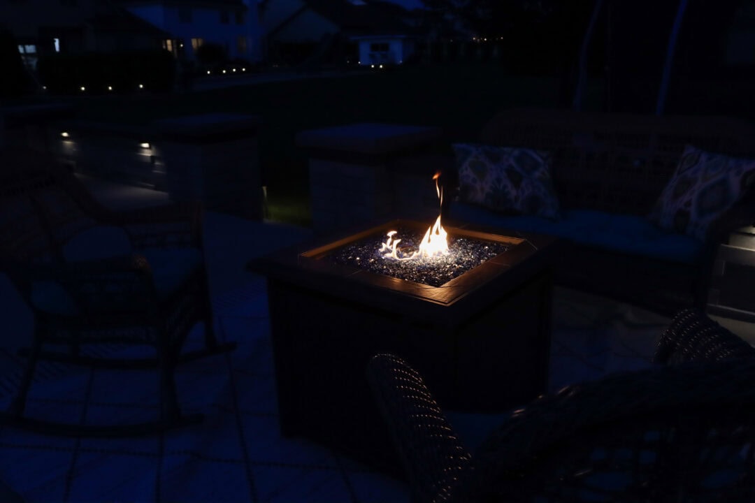 Landscape photo of a backyard at night featuring a fire pit near the center of the image. Chairs can be seen surrounding the fire pit. Small lights can be seen in the surrounding area.