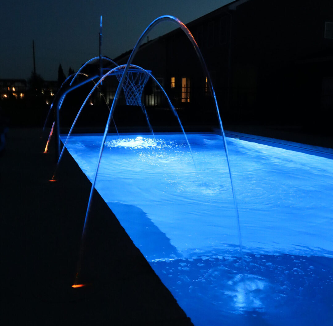 Square image of four laminars shooting curved, solid arcs of water into a glowing blue inground fiberglass pool at nighttime. A basketball hoop is placed in the middle of the four laminars on the left side of the pool/image. Beyond the pool is black, blurred houses, the lights from which can be seen glowing in the background.