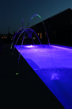 Portrait photo of a glowing purple inground pool featuring three glowing laminars arcing water into the pool from the left side of the image.