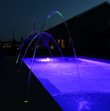 Portrait photo of a glowing purple inground pool featuring three glowing laminars arcing water into the pool from the left side of the image.