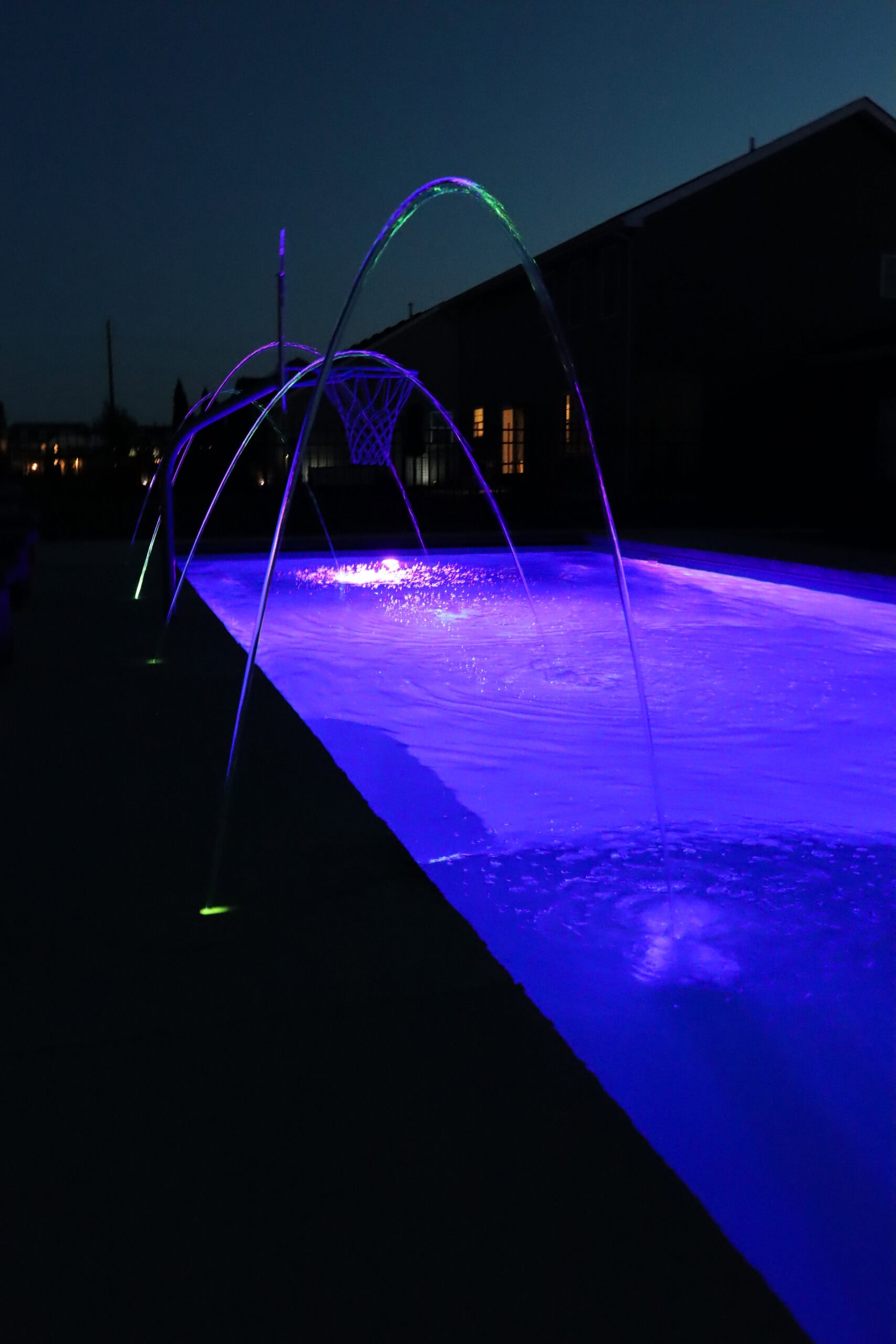 Portrait photo of laminars shooting arches of water into a purple glowing inground pool at night.