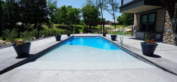 Image taken from the front of a light-blue, beach-entry fiberglass pool surrounded by a concrete pool deck off the back of a house. The pool is surrounded by six potted plants, three along both lengths. Trees and shrubbery can be seen in the backyard behind the pool.