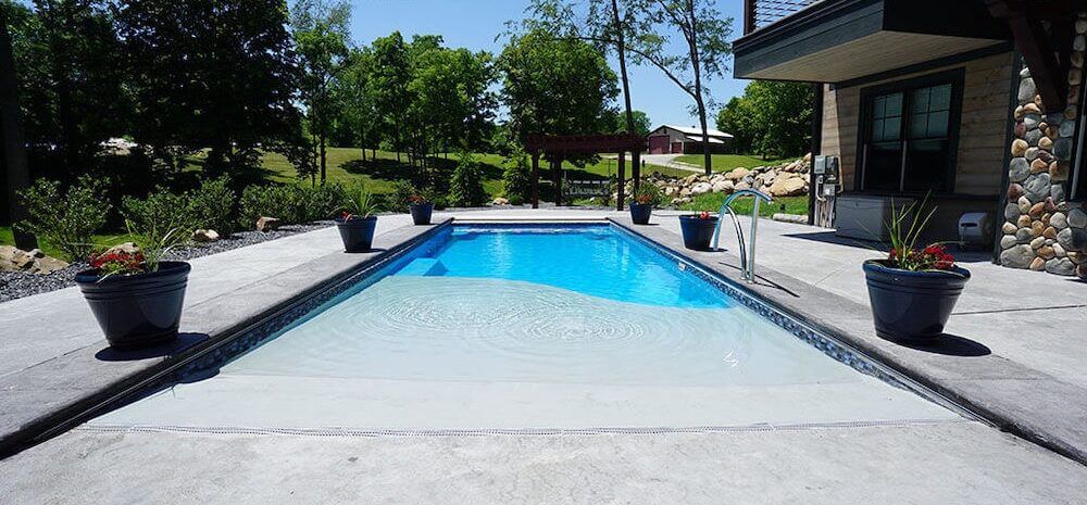 Image taken from the front of a light-blue, beach-entry fiberglass pool surrounded by a concrete pool deck off the back of a house. The pool is surrounded by six potted plants, three along both lengths. Trees and shrubbery can be seen in the backyard behind the pool.