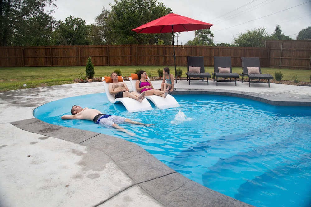 Photo of three people resting on the tanning ledge of a sandal beach-entry fiberglass pool from Thursday Pools. Behind them are lounge chairs, shrubs, and fence encasing the backyard.