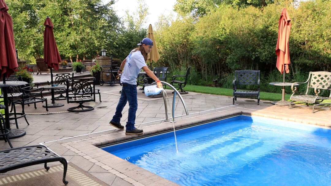 Photo of a man standing over the shallow end of a light-blue inground pool pouring a yellow liquid into the pool. Behind the pool is a stamped concrete patio with chairs and a retaining wall. Trees can be seen behind the patio.