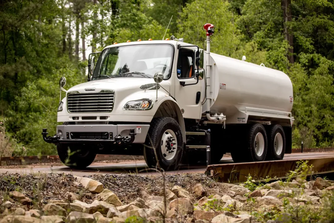 Landscape photo of a white water truck parked on a bridge in the middle of a forest. Rocks can be seen in the foreground in front of the truck and bridge.