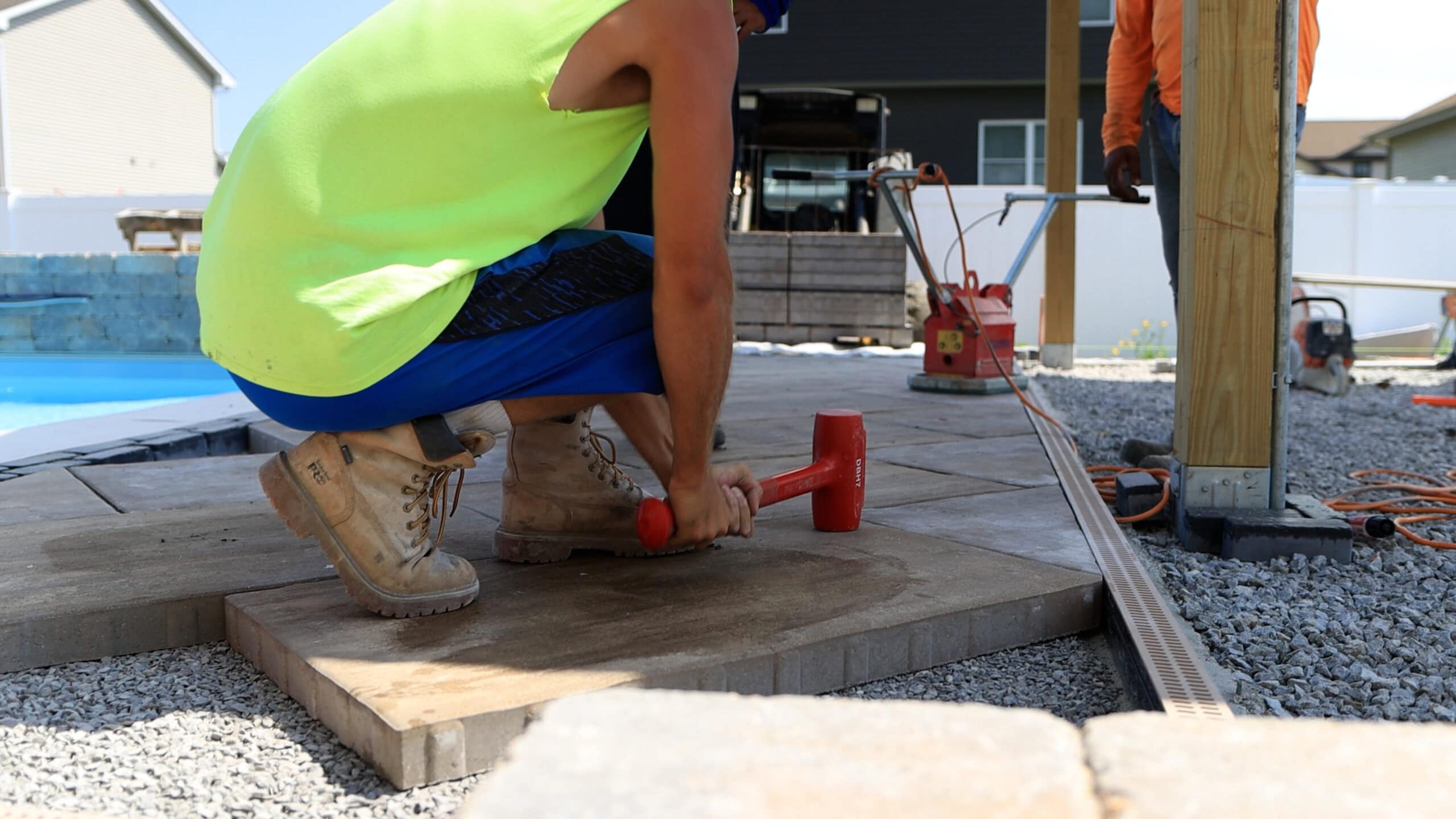 Man wearing a neon yellow tank top and blue shorts crouched down pounding a large concrete paver into place over a gravel bed.