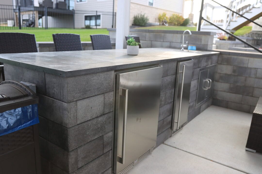 Landscape photo of the counter of an outdoor kitchen. Underneath the counter are two stainless steel cabinets. Atop the counter on the far side near the background is a sink. Cut off by the left side of the image in the foreground is a garbage can. Three chairs can be seen on the other side of the counter. A small potted plant also sits in the middle of the counter. Grass and the bottoms of houses can be seen in the background.