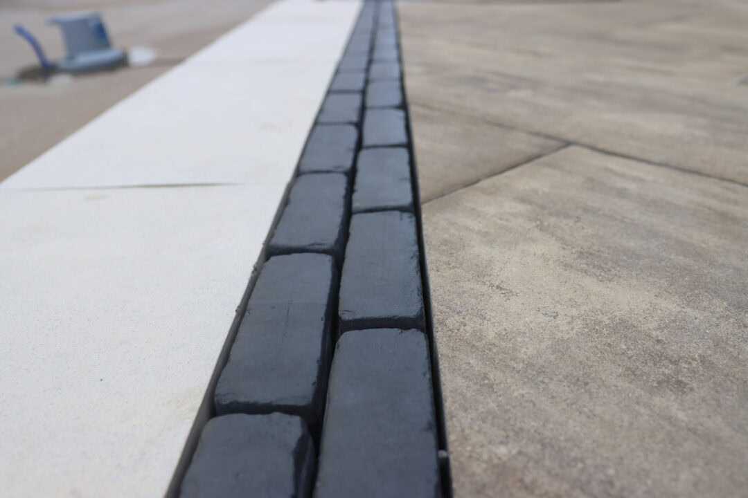 Landscape photo taken up close of a black brick paver patio border running parallel along a light gray inground pool concrete border. The paver border is skinny and runs slightly left of center of the image. The pool's automatic cover is visible in the top left corner of the image. Larger gray pavers are visible right of the black pavers.