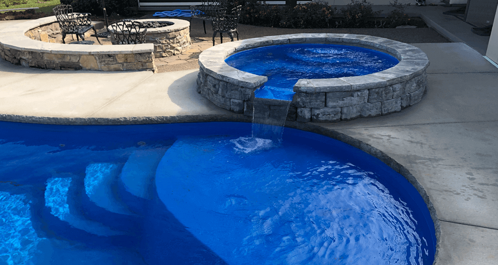 Tanning ledge and steps of a blue freeform fiberglass inground pool with a spillover spa near the tanning ledge spilling water into the pool. In the background is a fireplace surrounded by chairs.