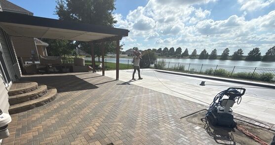 Partially complete brick paver patio off the back of a house with a covered inground pool. In the background is a pond with trees behind it.