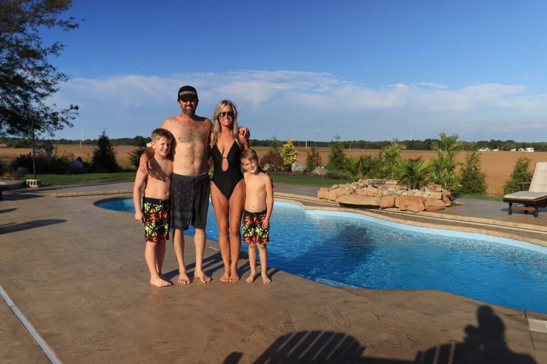 Landscape photo of a family of four standing in front of a blue, freeform inground pool, surrounded by a large concrete patio. The family is just left of center of the image, casting a shadow over the middle of the pool. Beyond the pool patio are trees, grass, and an empty cornfield. the sky takes up the top third of the photo. Light clouds can be seen on the horizon. 