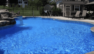 Landscape photo taken at a downward-angle towards a large freeform vinyl liner pool that takes up the bottom two-thirds of the image. An overhanging rock waterfall can be seen on the left-side of the image. The pool features a basketball hoop in the background off the pool's shallow end. In the upper right corner of the image is the back of a house featuring some bushes outside the windows. A black fence runs along the top left side of the image behind the pool and house. Grass can be seen beyond the fence.