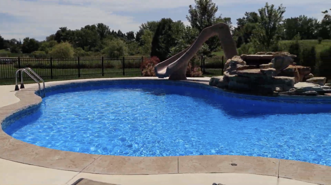 Landscape photo of a portion of a backyard pool and deck. The blue freeform pool runs across the bottom two-thirds of the image. It is wider on the left side and curves inward in the center and runs to the edge of the right-side of the photo. On the right side of the image on the other side of the pool is a slide and rock waterfall. Beyond the pool, in the background is grass and a lack fence. More grass, trees, and shrubbery sit beyond the fence in the background. The sky can be seen at the top of the image.