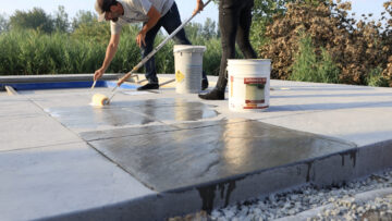 Landscape photo taken from beyond the edge of a stamped concrete pool patio. A man and woman stand on the concrete in the middle of the image. The woman is rolling a wet substance onto the concrete. The slab closest to the camera to her is all wet. The man leans down and holds his hand close to the concrete. In the background is tall grass and shrubbery. The sky can be seen above the trees. Two buckets sit on the concrete between the man and the woman.