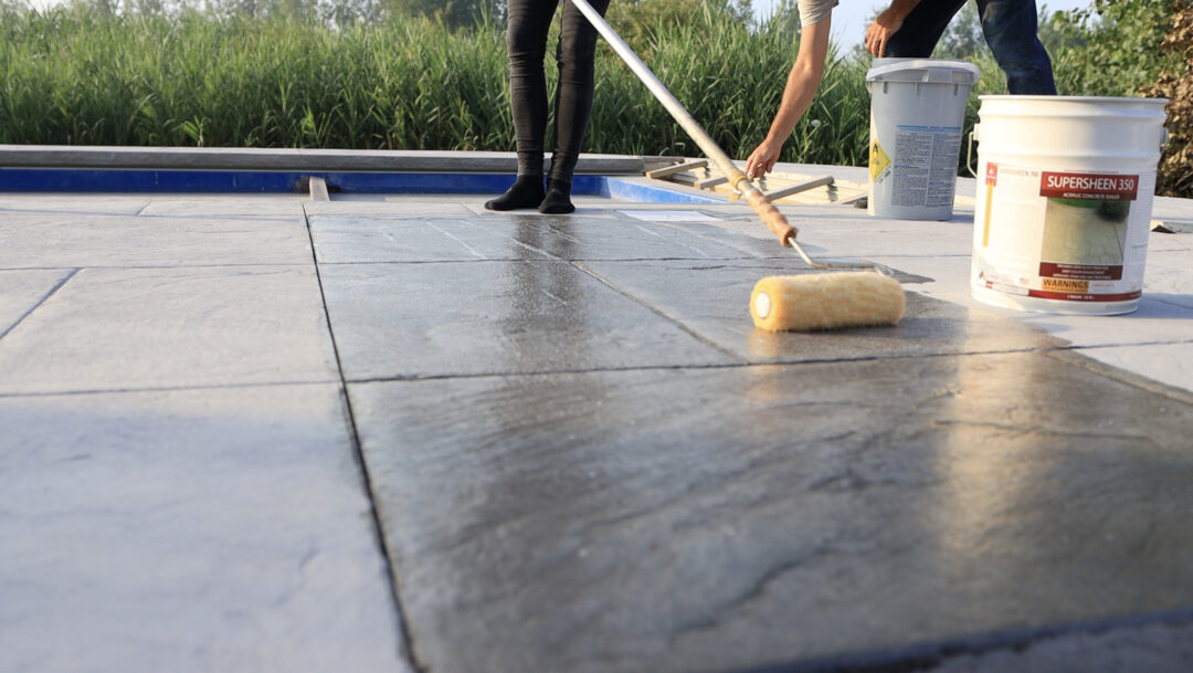 Landscape photo of a stamped concrete pool patio being sealed with a roller. Two pairs of legs can be seen standing on the concrete in the top right corner of the image with two buckets sitting near them. In the background is tall grass.