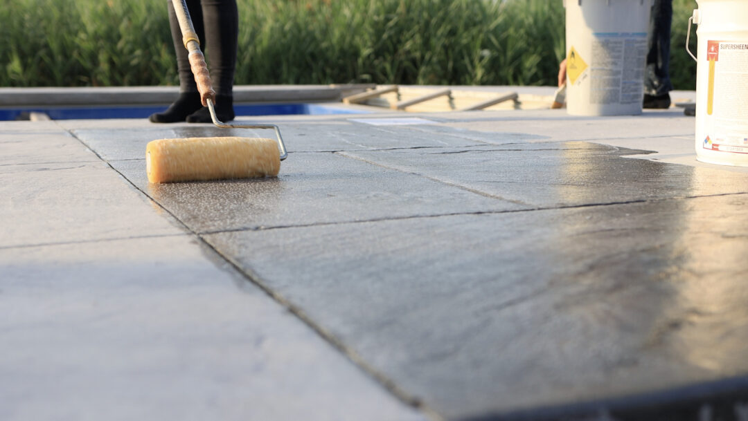 Landscape photo taken near ground level of a partially-wet stamped concrete pool patio. Left of center at the top of the image is a pair of legs and feet standing on the concrete. From that spot extends a pole with a fabric roller rolling a substance onto the concrete. At the top of the image in the background is tall grass.