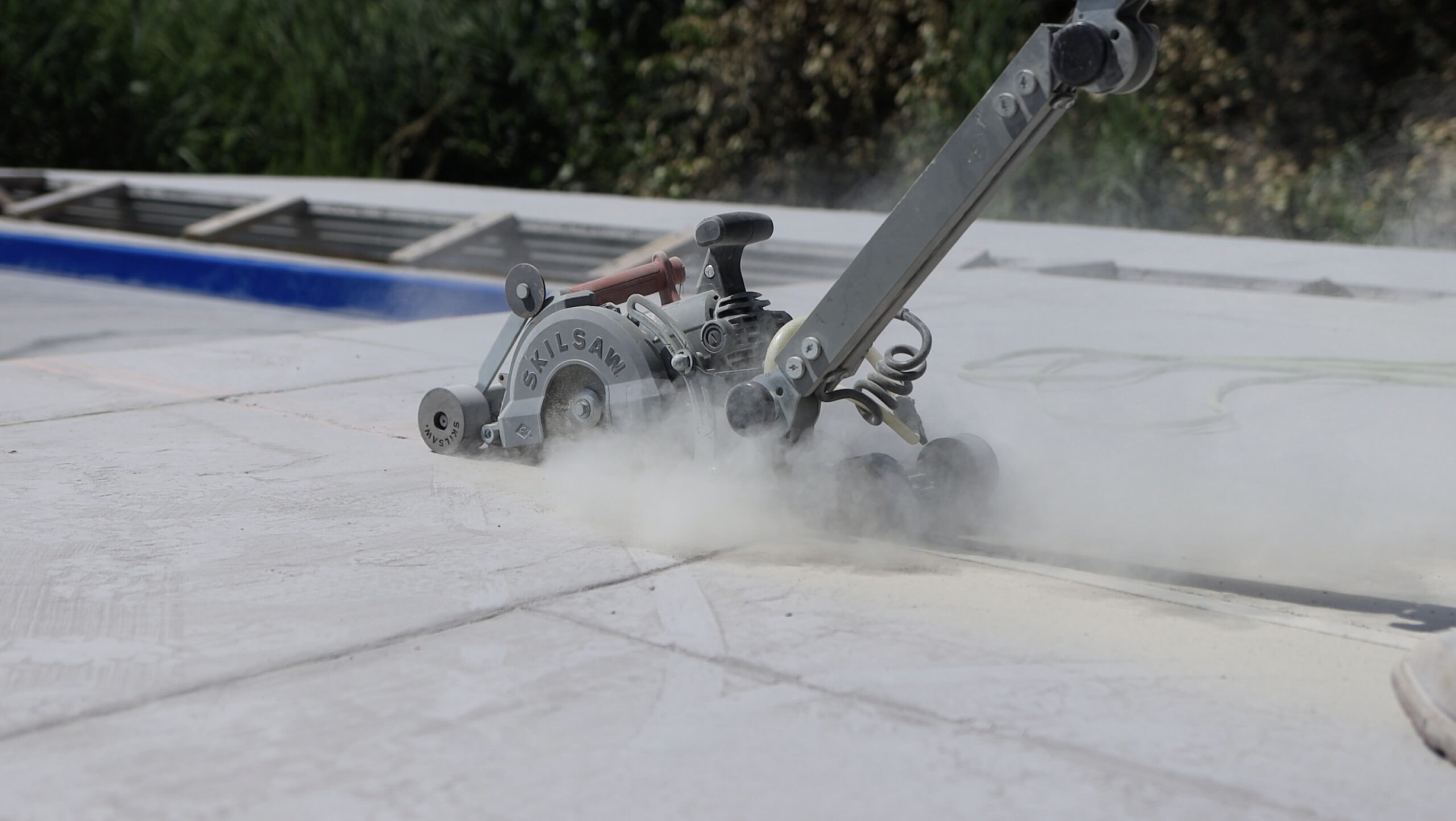 Landscape photo of a concrete cutter cutting joints into a stamped concrete pool patio. The machine is in the middle of the image and sending dust up around its blade. The pool can be seen partially on the left side of the image. At the top of the image in the background is tall grass and shrubbery.